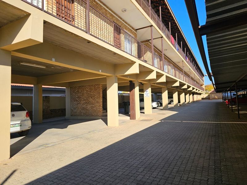 Exterior view of an apartment complex featuring a paved driveway with covered parking bays and multi-level balconies with metal railings.