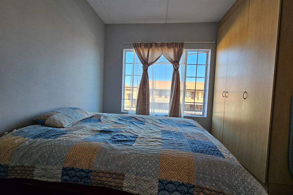 A functional bedroom featuring built-in wooden cupboards and a window with security bars, offering natural light and views of the surrounding complex.