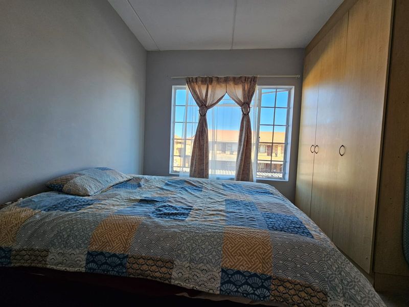 A functional bedroom featuring built-in wooden cupboards and a window with security bars, offering natural light and views of the surrounding complex.