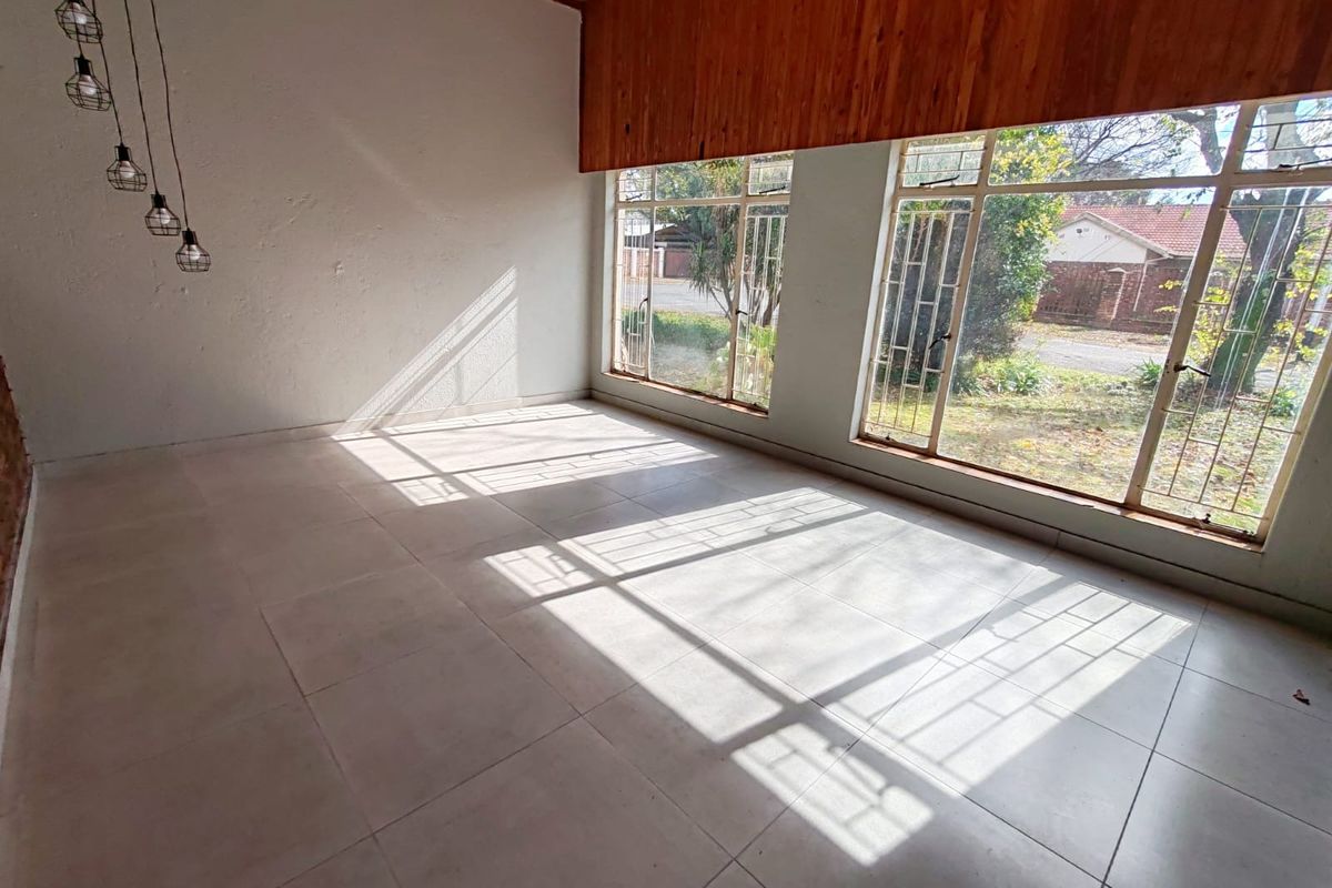 A bright, empty living room featuring large windows with security bars, tiled flooring, and a distinctive timber ceiling accent.