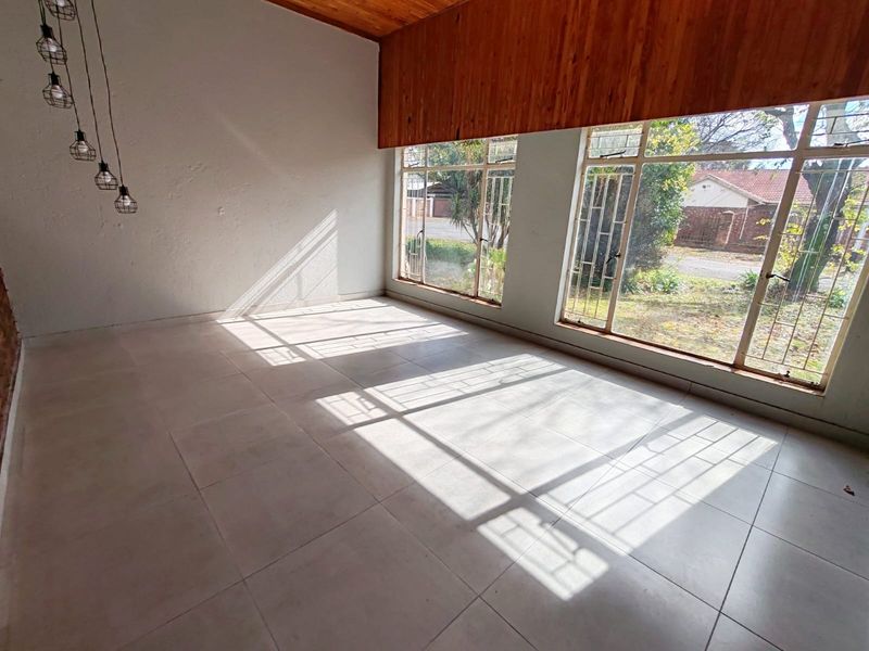 A bright, empty living room featuring large windows with security bars, tiled flooring, and a distinctive timber ceiling accent.