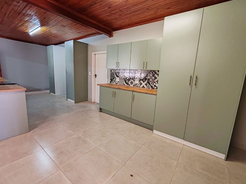 A modern kitchen featuring sage green cabinetry, wooden countertops, a decorative patterned tile backsplash, and a warm exposed timber ceiling.