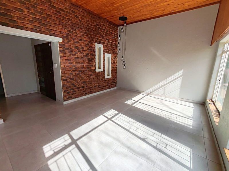 A bright, empty living room featuring an exposed brick accent wall, warm timber ceiling, and large windows that provide ample natural light.