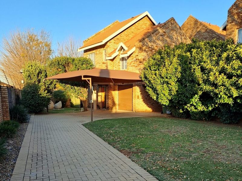 Front view of a brick townhouse featuring a paved driveway, a shadeport, a garage, and a well-maintained garden.