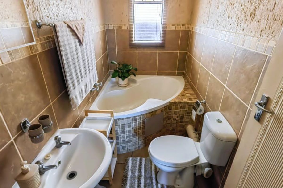 A compact bathroom featuring a corner bathtub, pedestal basin, and toilet with neutral-toned wall and floor tiling.