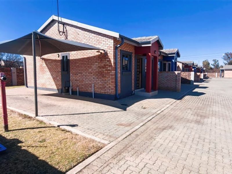 Exterior view of a face brick apartment unit featuring a paved driveway, shade cloth carport, and surrounding perimeter walling.