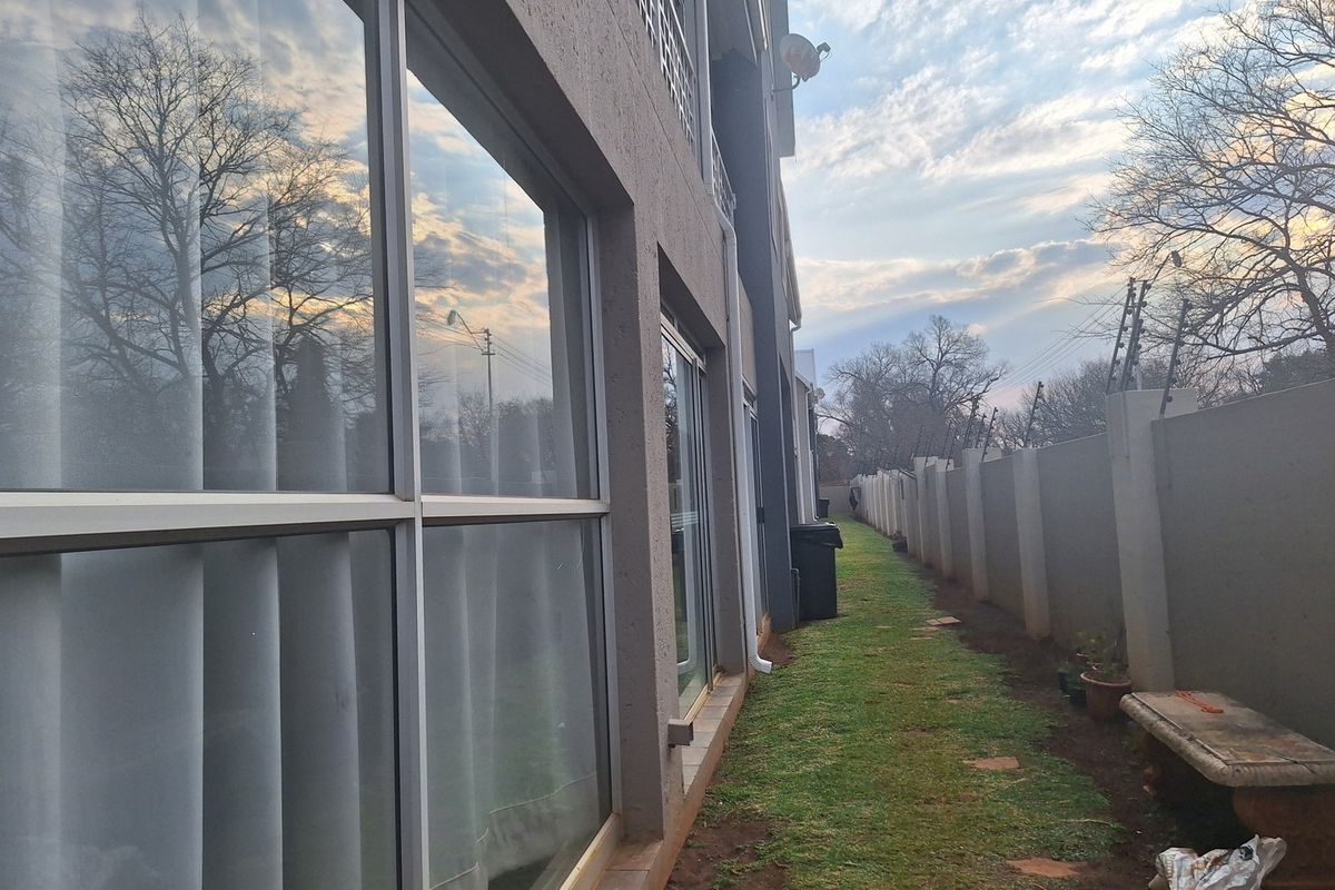 Rear exterior view of the apartment building featuring a narrow garden strip, perimeter wall with electric fencing, and large sliding glass doors.