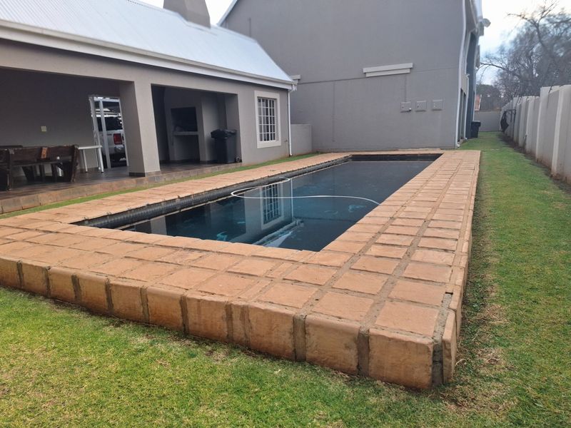 Outdoor pool area featuring a rectangular swimming pool with brick coping, adjacent to a covered patio with a built-in braai and garden space.