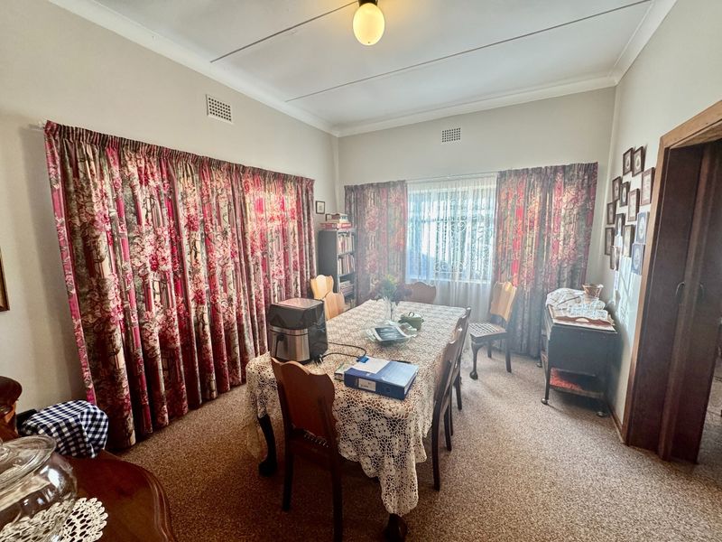 A traditional dining room featuring carpeted flooring, ceiling cornices, and large windows with floor-to-ceiling curtains.