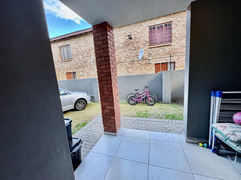 A covered patio area featuring tiled flooring and a structural brick pillar, overlooking a small paved courtyard and garden space.