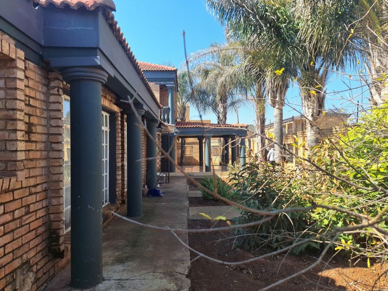 Rear exterior view of a brick house featuring a covered patio, concrete columns, and a paved walkway leading through the garden area.