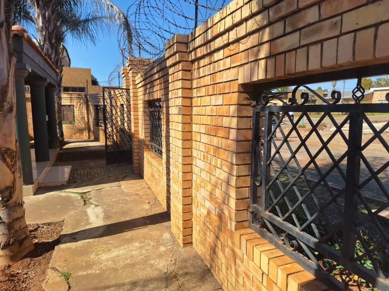 Front exterior view featuring a face brick boundary wall with wrought iron security gates, razor wire, and cracked concrete paving leading to a covered porch.