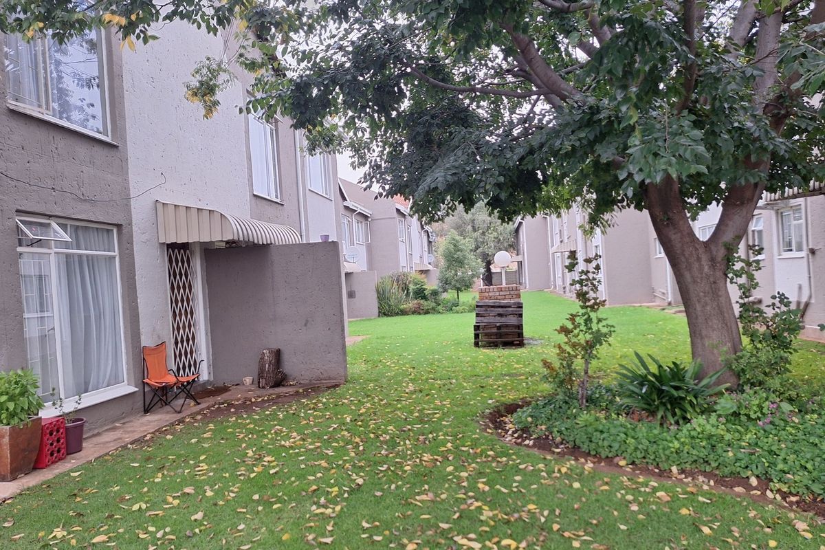 Rear view of the apartment complex featuring a shared garden area, mature trees, and private patio access with security gates.