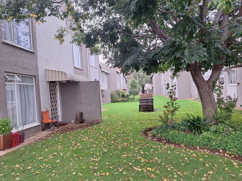 Rear view of the apartment complex featuring a shared garden area, mature trees, and private patio access with security gates.