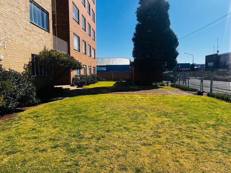 A view of the apartment building's rear exterior, featuring a brick facade, a well-maintained lawn, and a paved walkway enclosed by metal fencing.
