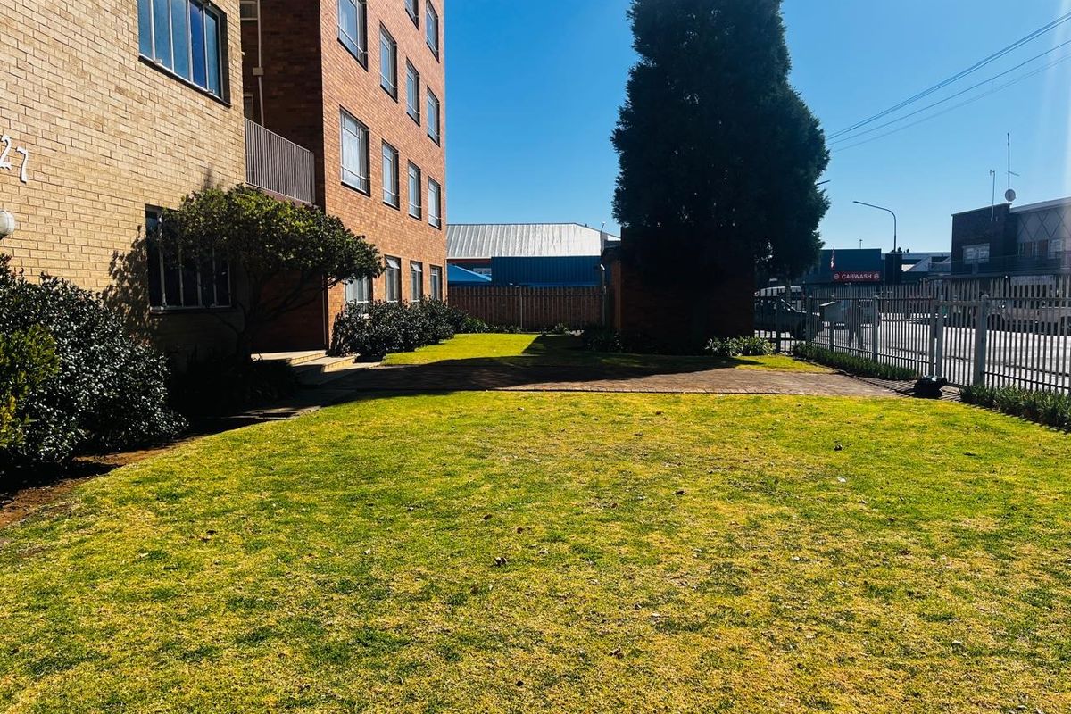 A view of the apartment building's rear exterior, featuring a brick facade, a well-maintained lawn, and a paved walkway enclosed by metal fencing.