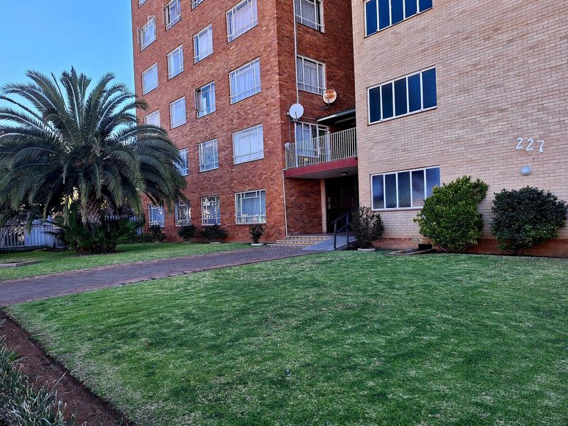 Front exterior view of a multi-story brick apartment building featuring a paved walkway, landscaped lawn, and a balcony entrance.