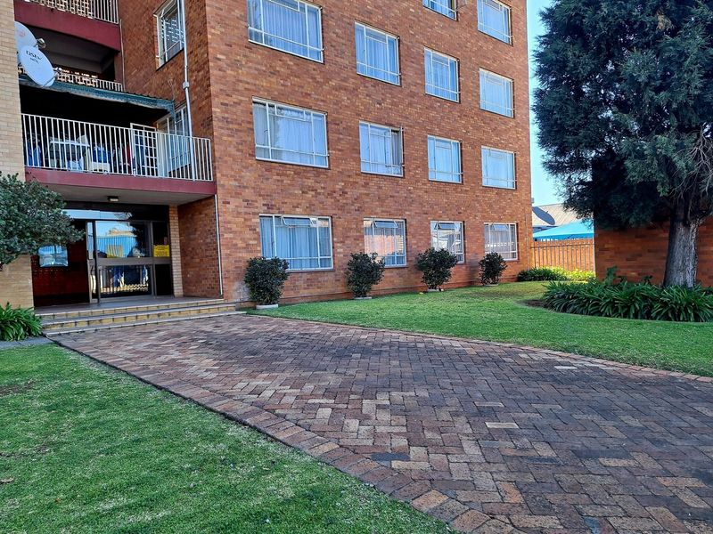 Exterior view of a multi-story brick apartment building featuring a paved driveway, landscaped garden, and balcony access.