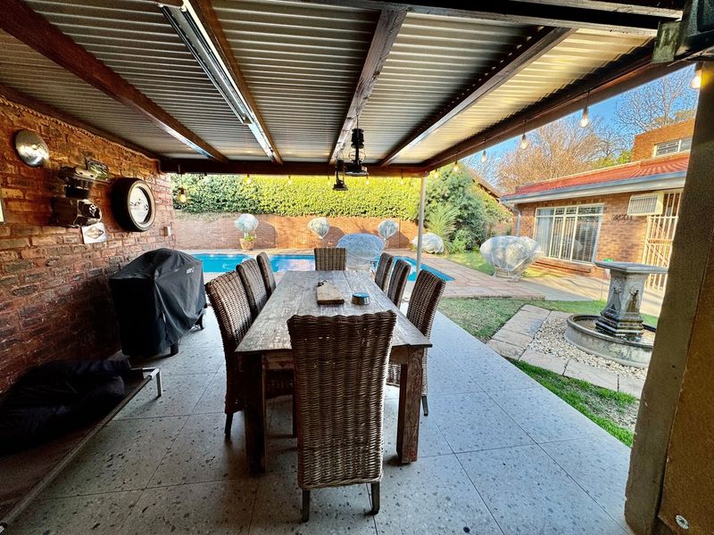 Covered outdoor patio area featuring tiled flooring, exposed brickwork, and a corrugated roof, overlooking a private swimming pool and garden water feature.