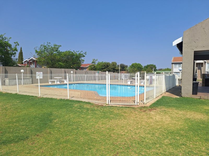 A communal swimming pool area featuring a paved surround, secure metal fencing, and a well-maintained lawn in a residential apartment complex.