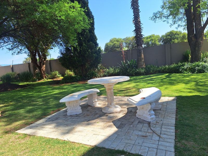 A well-maintained garden featuring a paved seating area with stone benches and a table, surrounded by established trees and a perimeter wall.