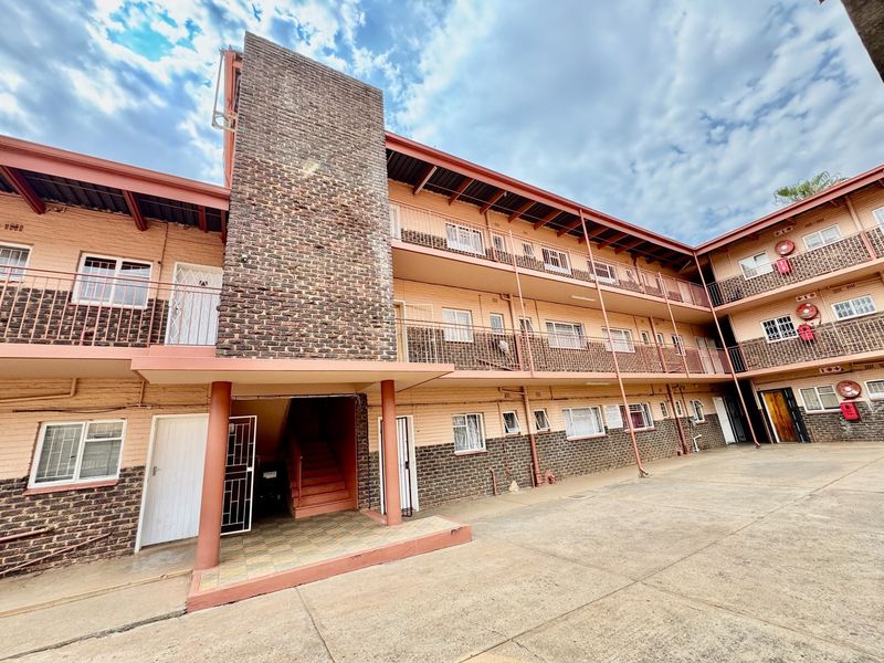 Exterior view of a multi-story apartment complex featuring brick facade, metal balcony railings, and a paved courtyard area.
