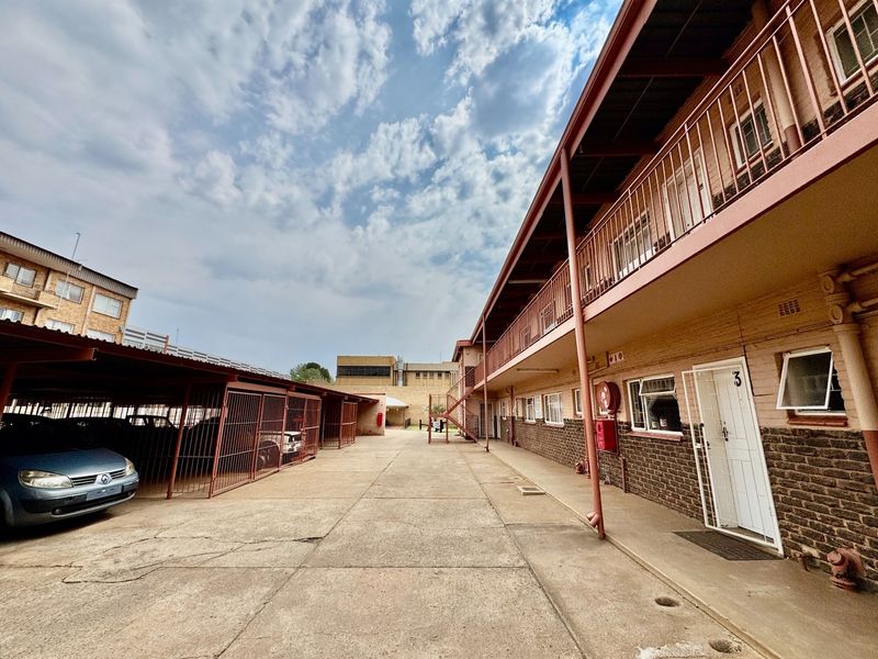 Exterior view of an apartment complex featuring covered parking bays, a concrete driveway, and access to ground floor units with security gates.