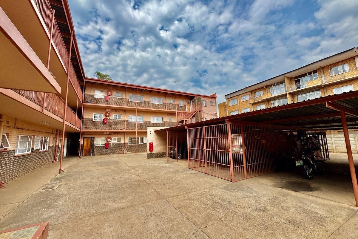 Exterior view of the apartment complex featuring a paved courtyard, covered parking bays with security cages, and multi-level access balconies.