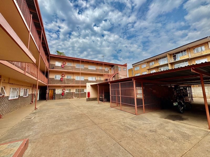 Exterior view of the apartment complex featuring a paved courtyard, covered parking bays with security cages, and multi-level access balconies.
