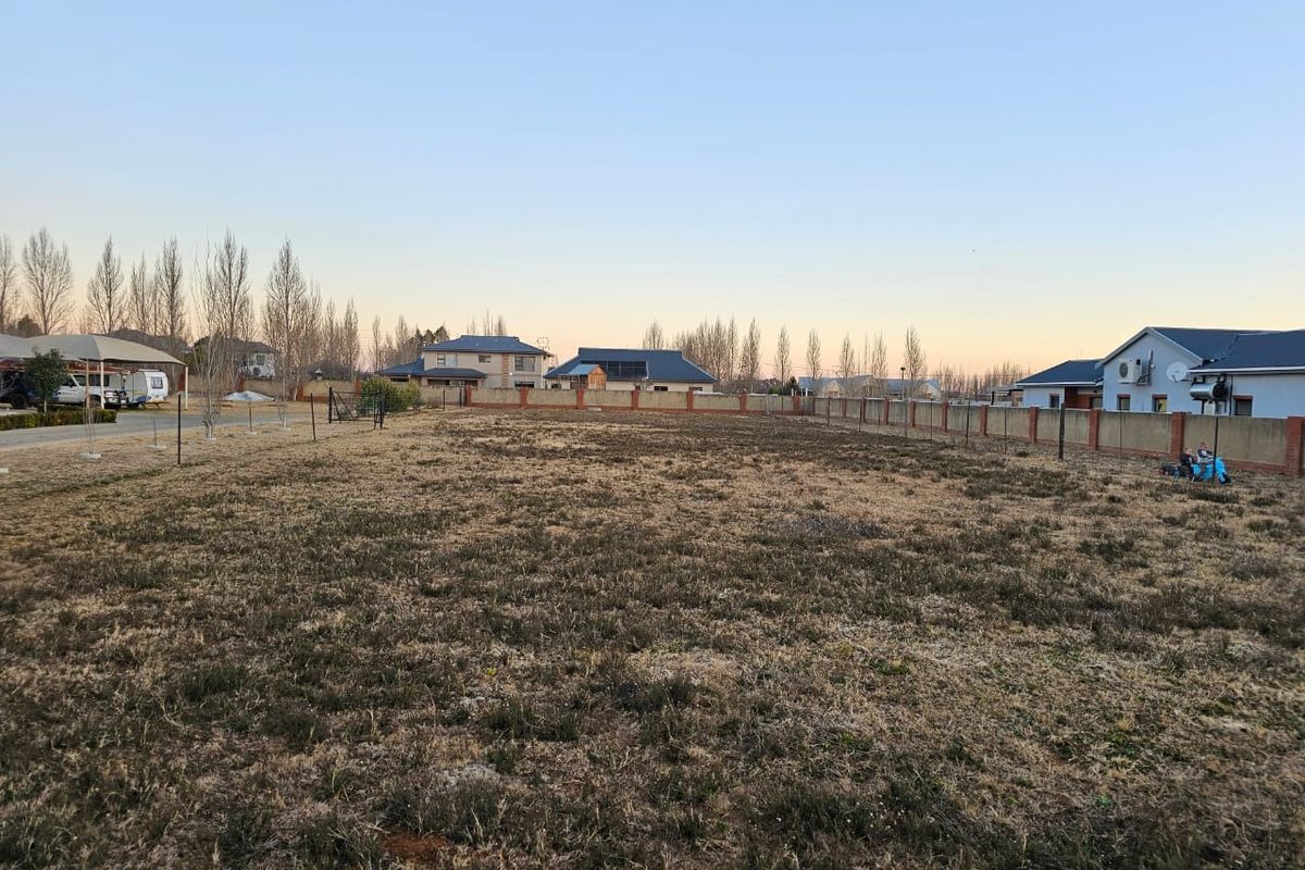 A vacant residential plot featuring dry grass, enclosed by a perimeter wall, situated in a developed neighborhood.