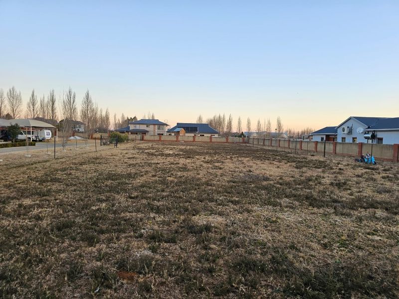 A vacant residential plot featuring dry grass, enclosed by a perimeter wall, situated in a developed neighborhood.