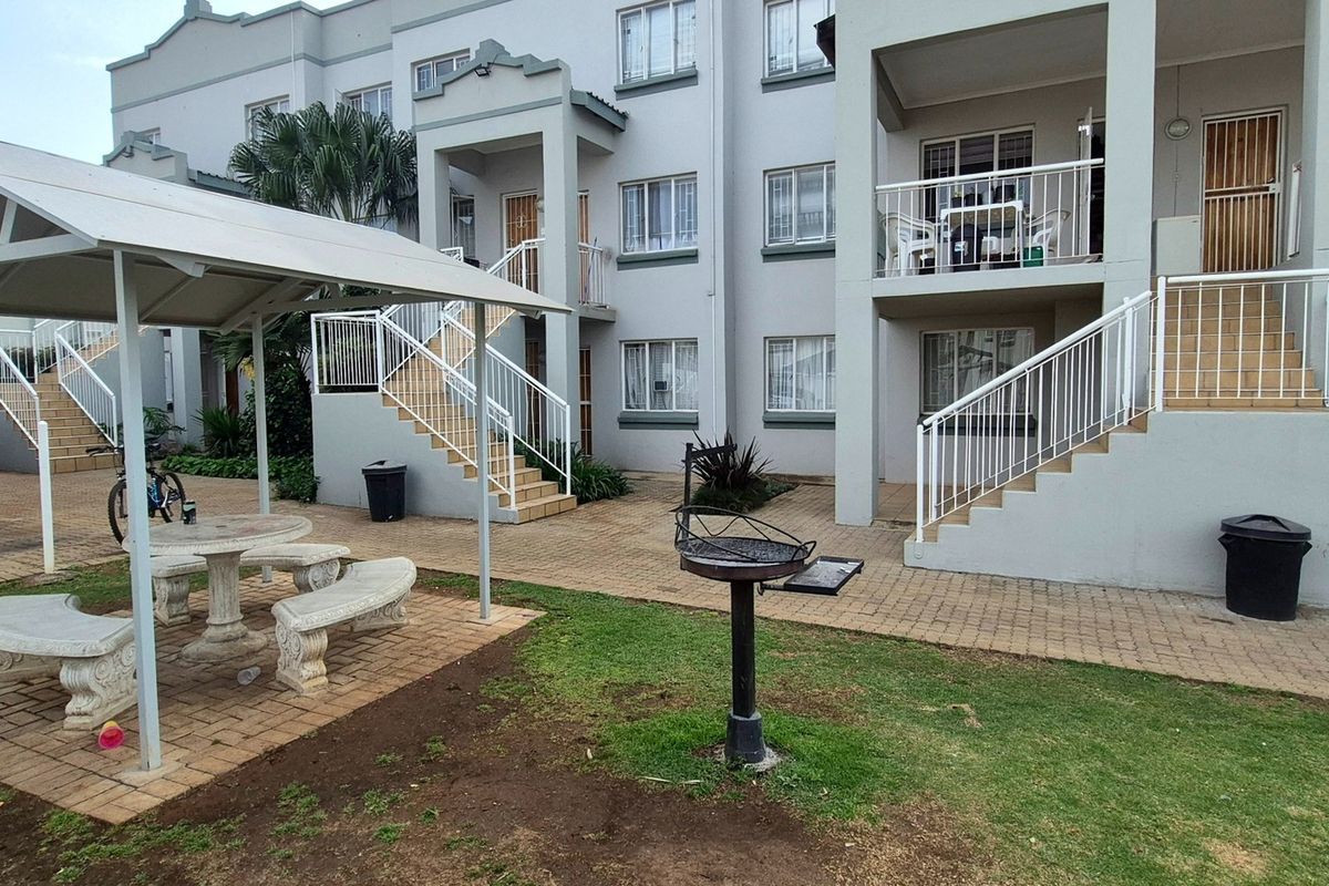 Exterior view of an apartment complex featuring a communal paved courtyard, covered seating area, braai facilities, and access staircases to upper-level units.