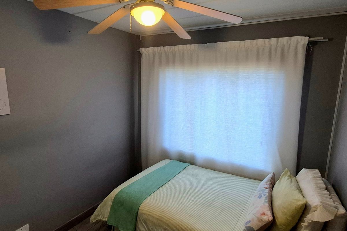 A compact bedroom featuring a ceiling fan and window with curtain rod, finished with neutral grey wall paint.