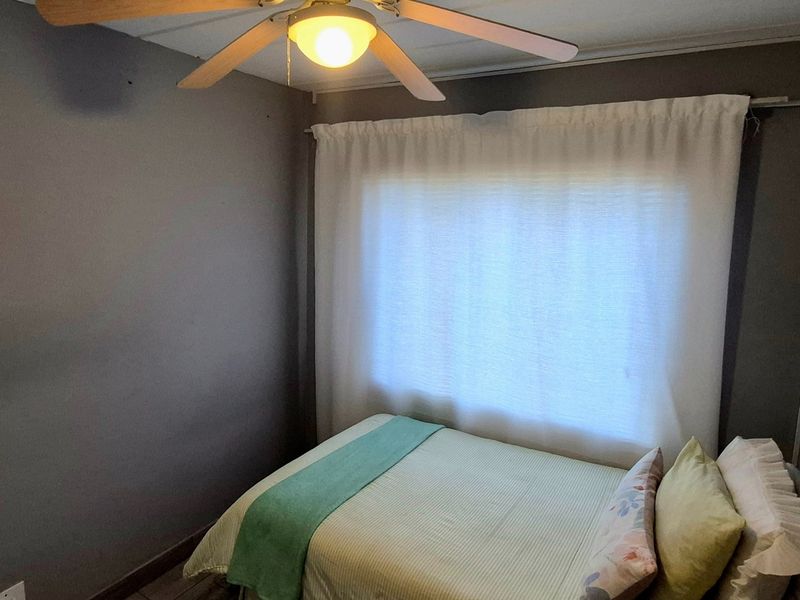 A compact bedroom featuring a ceiling fan and window with curtain rod, finished with neutral grey wall paint.