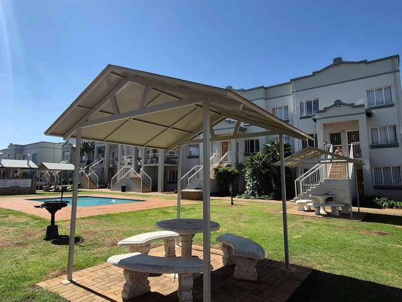 Communal outdoor area featuring a swimming pool, paved walkways, landscaped garden, and covered seating structures within an apartment complex.
