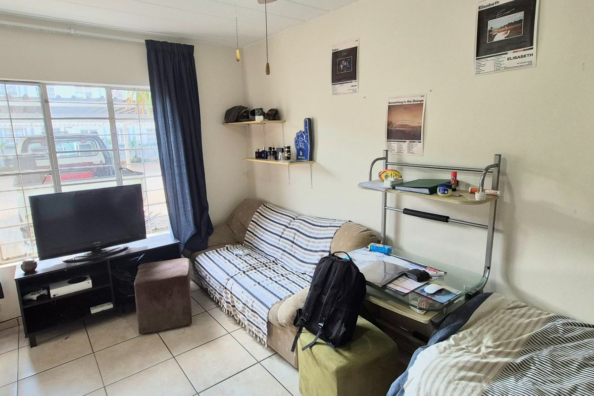 A functional bedroom featuring tiled flooring, a window with security bars, and a wooden door, providing a basic living space.