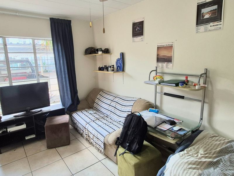 A functional bedroom featuring tiled flooring, a window with security bars, and a wooden door, providing a basic living space.