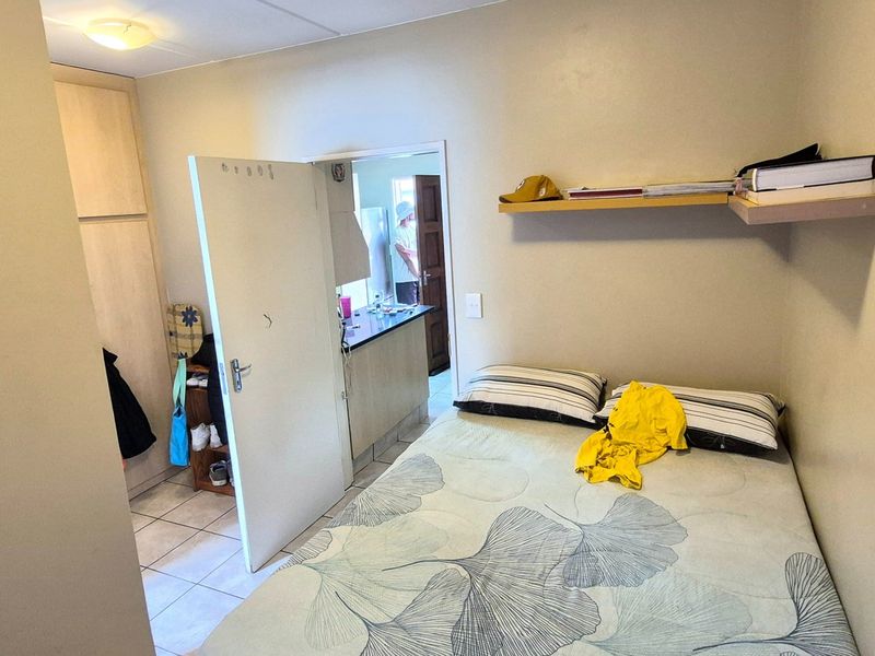 A compact bedroom featuring built-in cupboards, a floating wall shelf, and tiled flooring, with an adjacent view into the kitchen area.