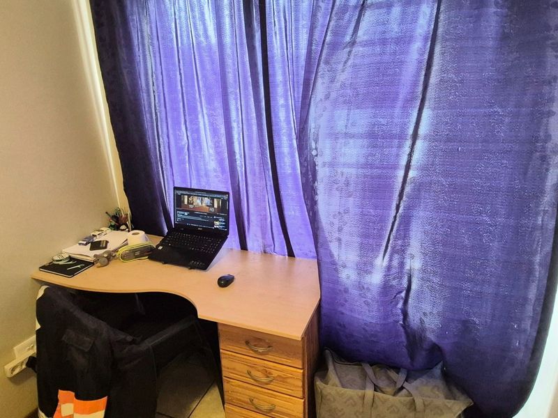 A small bedroom space featuring a window with curtains and an electrical outlet, currently utilized as a home office area.