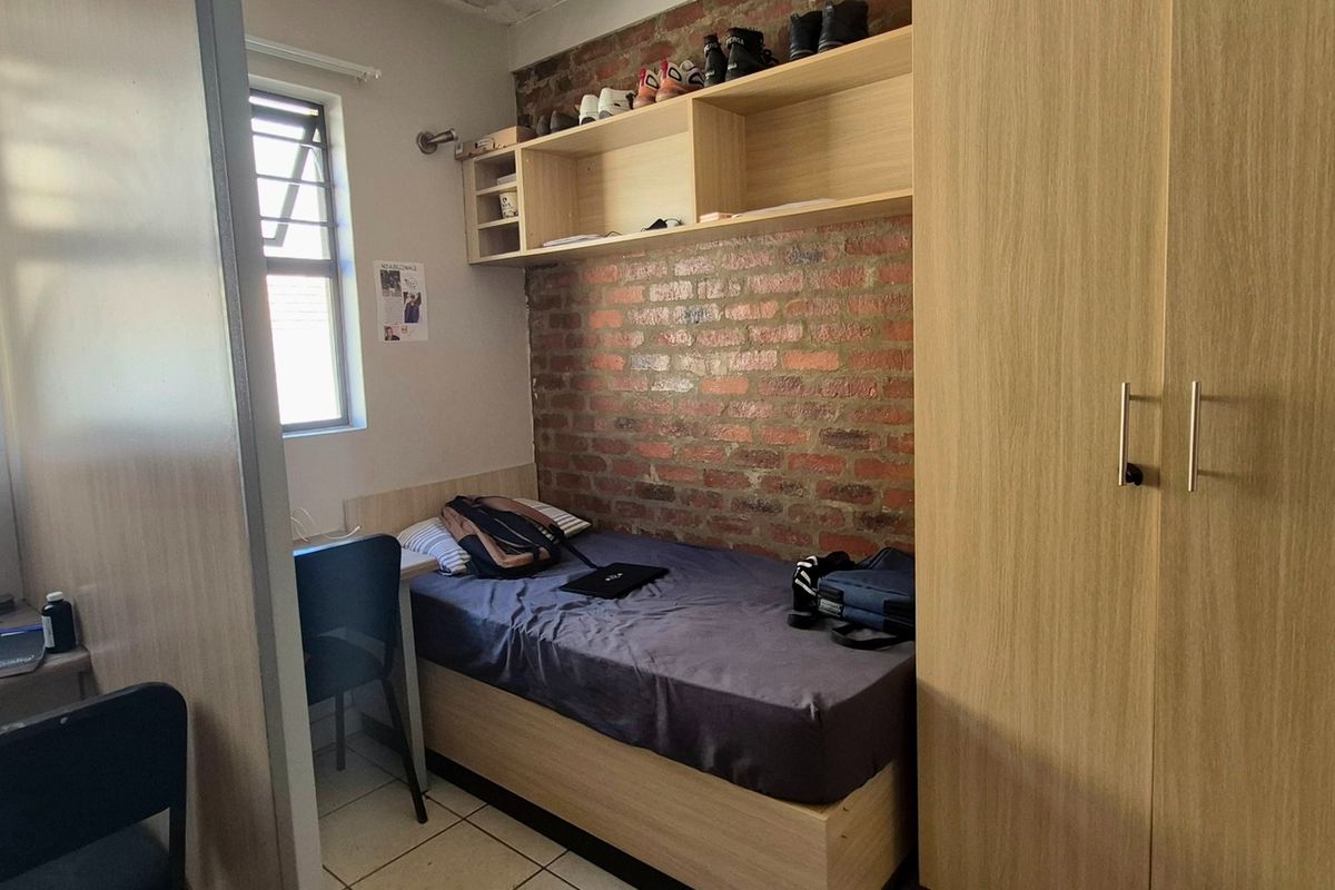 A compact bedroom featuring an exposed brick feature wall, built-in wardrobe, overhead shelving, and tiled flooring.
