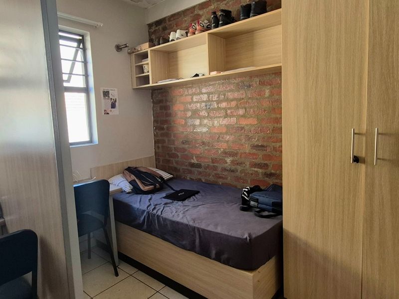 A compact bedroom featuring an exposed brick feature wall, built-in wardrobe, overhead shelving, and tiled flooring.