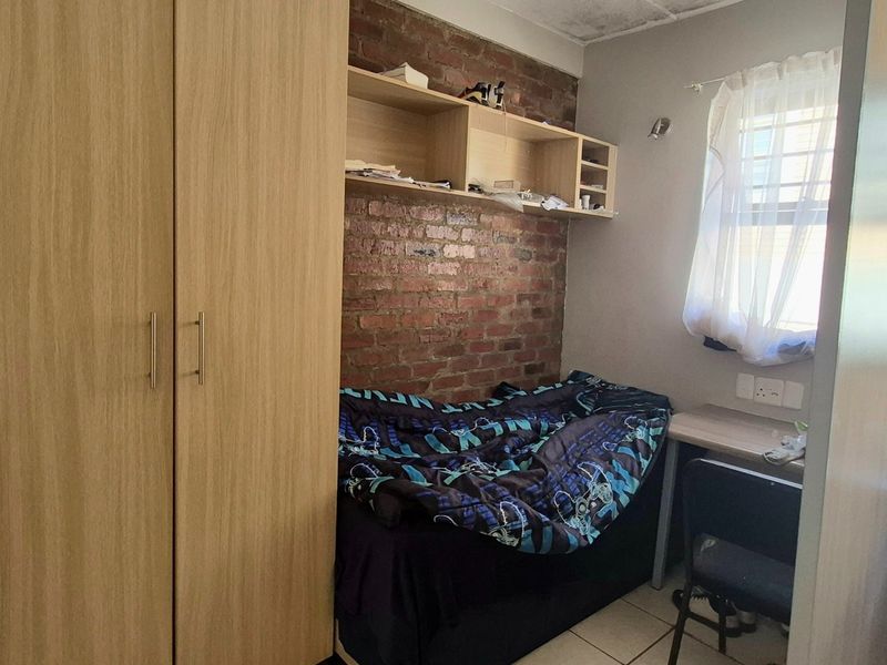Compact bedroom featuring a built-in wardrobe, exposed brick accent wall, integrated shelving, and a built-in desk area with tiled flooring.
