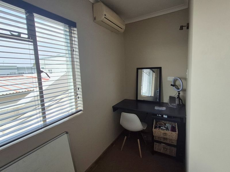 A compact home office featuring a built-in desk, wall-mounted air conditioning unit, and large window with venetian blinds.