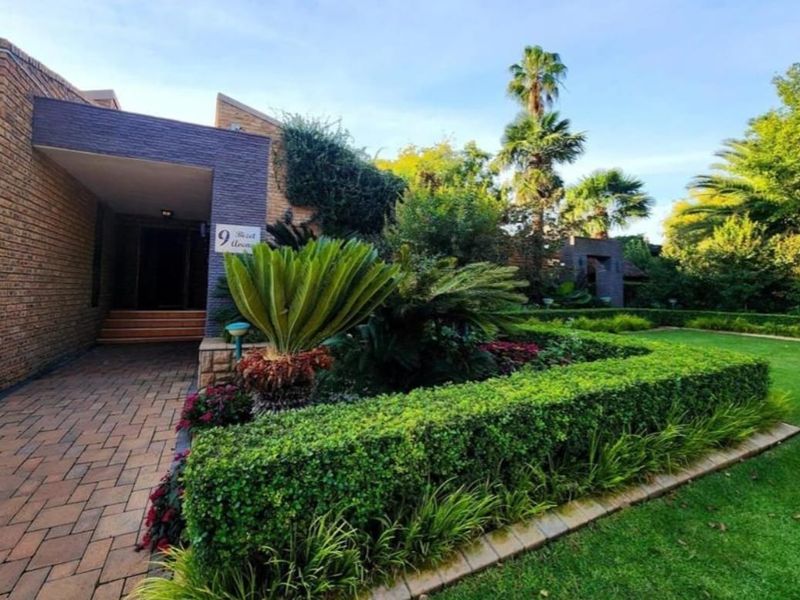 A well-maintained front entrance featuring a brick facade, paved walkway, and manicured garden with established hedges and lush landscaping.
