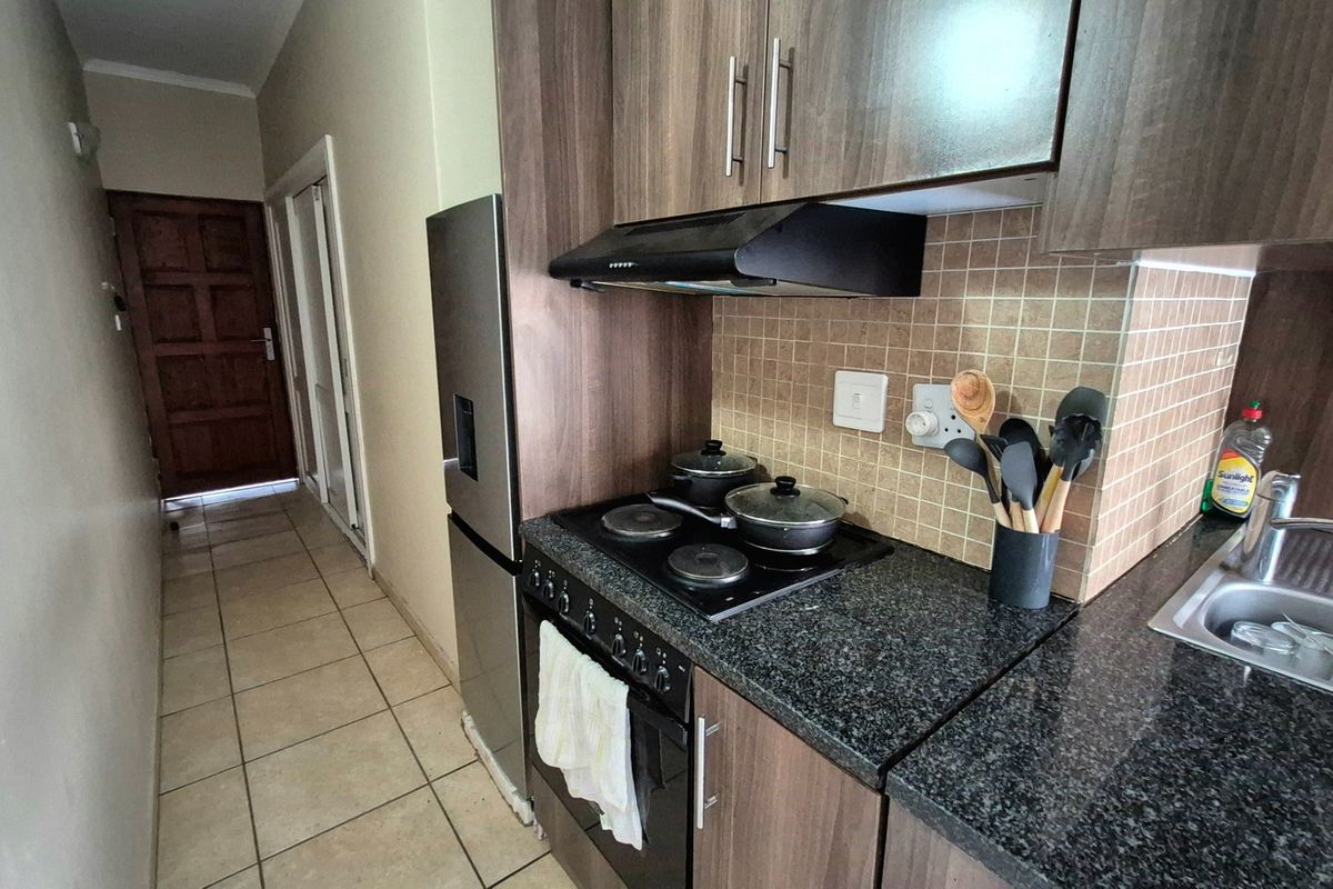 A compact kitchen featuring dark wood cabinetry, granite countertops, a built-in oven with hob, and a tiled backsplash, leading into a tiled hallway.