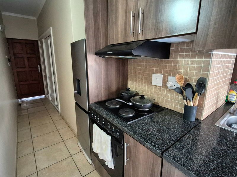 A compact kitchen featuring dark wood cabinetry, granite countertops, a built-in oven with hob, and a tiled backsplash, leading into a tiled hallway.
