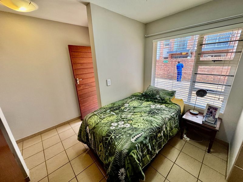 A functional bedroom featuring tiled flooring, a timber door, and a large window with horizontal blinds, providing natural light.