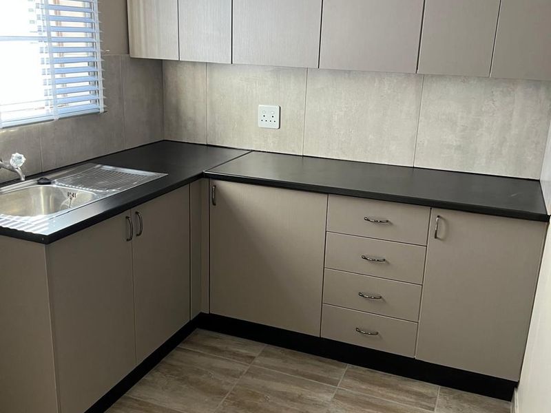 A clean and functional kitchen featuring modern l-shaped cabinetry, dark laminate countertops, a stainless steel sink, and neutral tiled flooring.