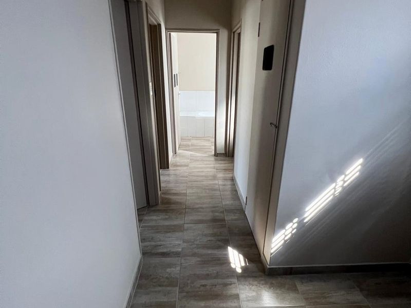 A clean, well-lit hallway featuring neutral tiled flooring and access to multiple rooms, including a glimpse of a bathroom at the end.