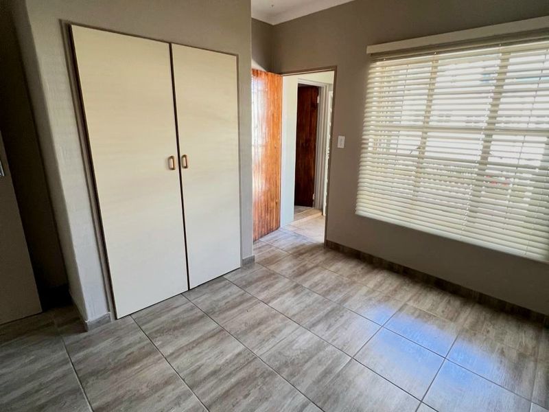 A clean, empty bedroom featuring neutral wall tones, tiled flooring, a built-in wardrobe, and venetian blinds on the window.